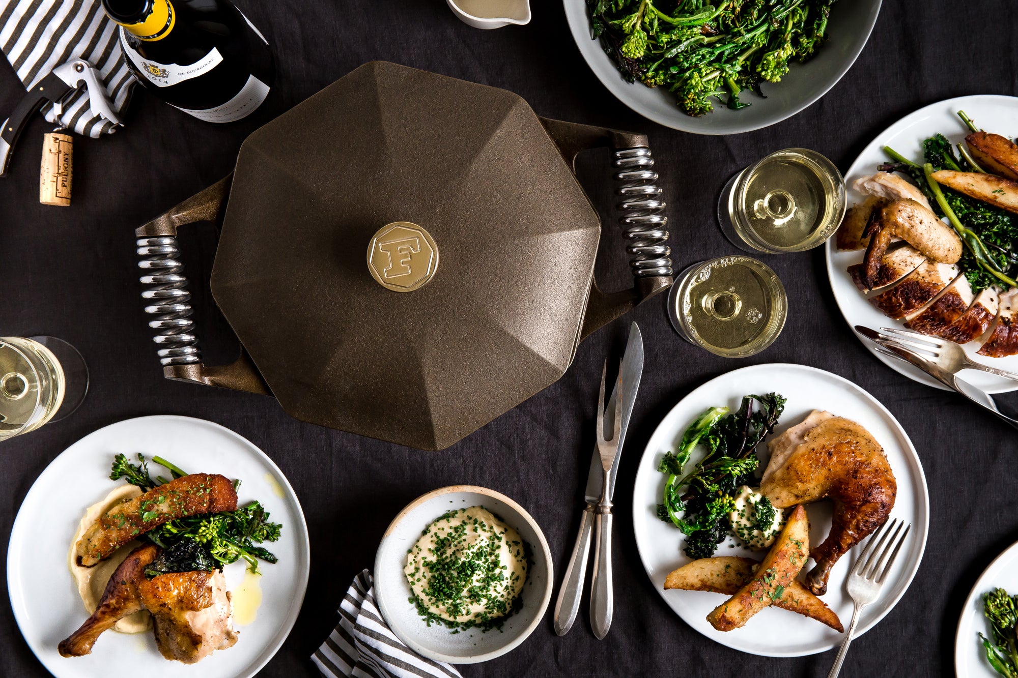 An overhead dining table scene featuring a large dark brown geometric lidded pot or casserole dish at the center, surrounded by plated roasted chicken, broccoli, mashed potatoes, wine glasses, and cutlery on a black tablecloth.